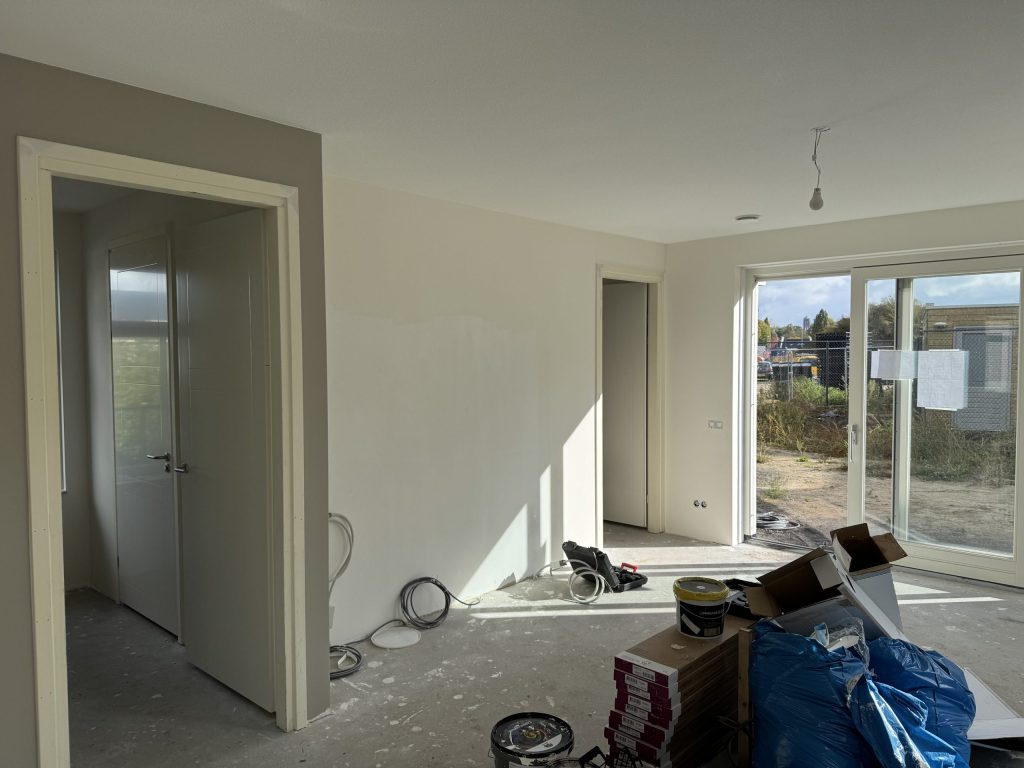 Woonkamer van een nieuwbouwwoning tijdens de afwerking, met verfemmers en dozen op de betonvloer