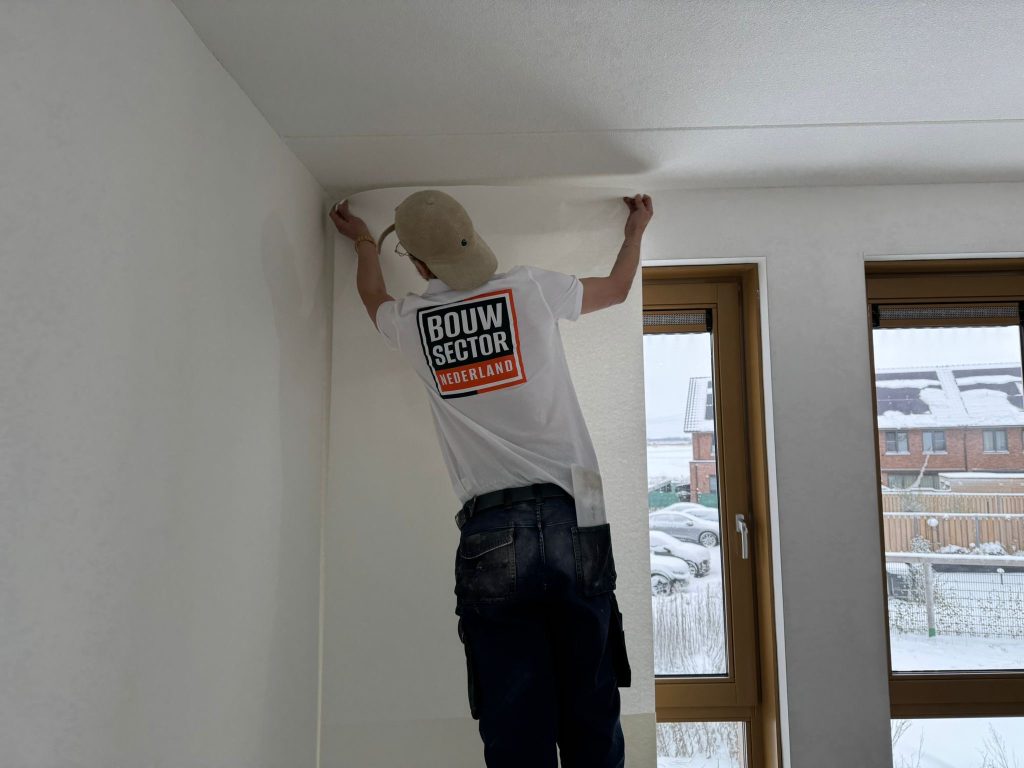 Behanger van Bouwsector Nederland zet een baan vlies vanaf het plafond tegen de muur van een nieuwbouwkamer
